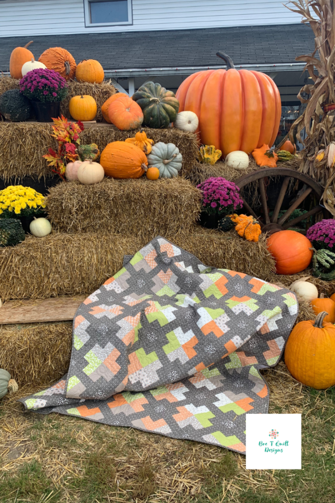 Finished fall jelly roll quilt with oranges, greens, and greys featured in front of a pumpkin fall display draped over straw bales