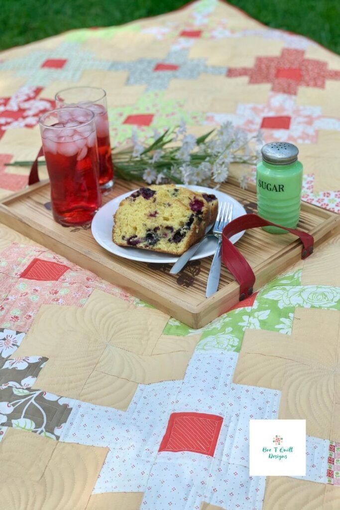 Yellow simple jelly roll quilt with platter of blueberry bread and red drinks on it like a picnic quilt. 